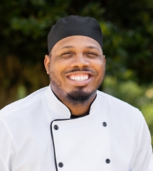 Chef Cyril (Cy) Stewart smiling in a white uniform and black hat outside a senior living unit.