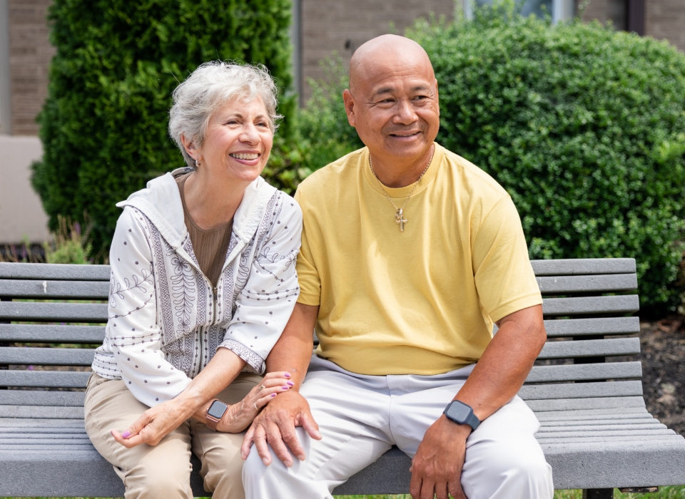 Two smiling seniors sitting on a bench, surrounded by greenery at a residential community.
