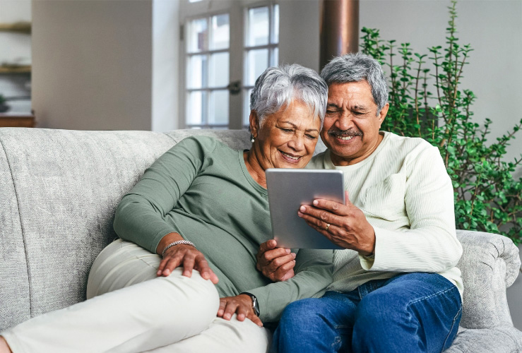 Senior couple sitting on a sofa smiling while looking at a tablet.
