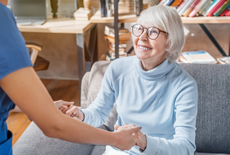 Smiling senior woman in glasses holding hands with a caregiver in a cozy living room